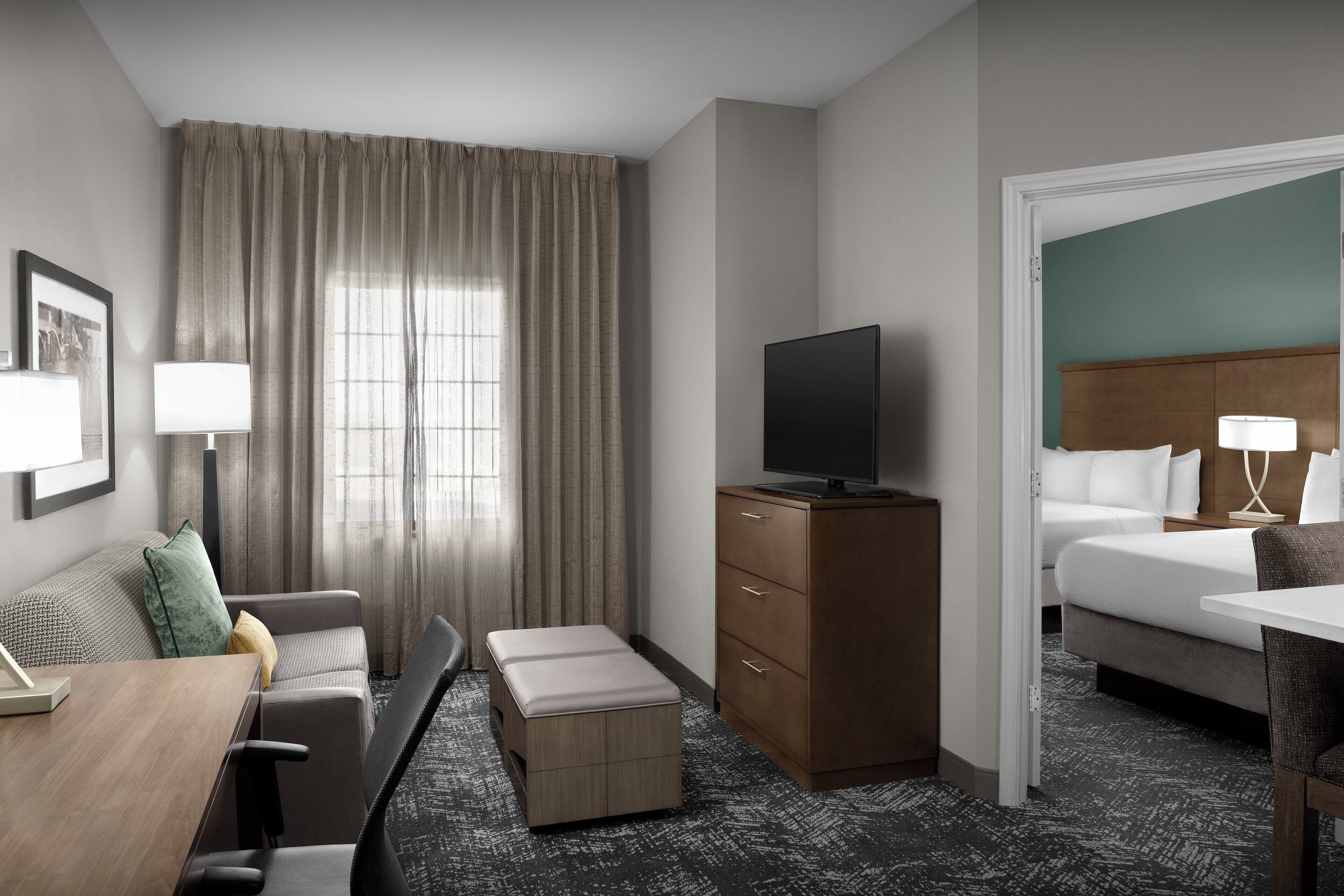 Une chambre d’hôtel avec un canapé, un bureau, un téléviseur posé sur un meuble en bois, un lit avec une tête de lit et une fenêtre avec des rideaux.