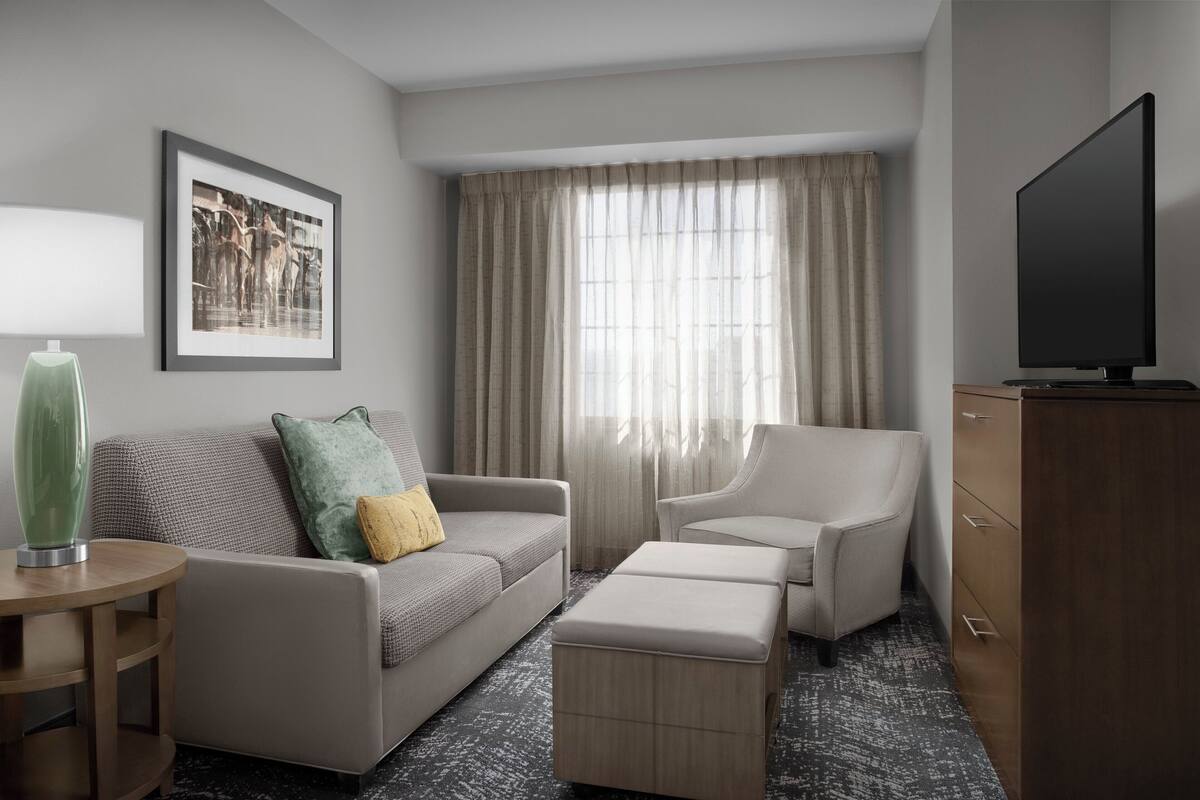 Une chambre d’hôtel moderne avec un canapé gris, un fauteuil assorti, une commode en bois et un téléviseur à écran plat.