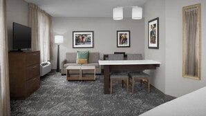 A hotel room with a flat-screen TV, a sofa with cushions, a dining table with chairs, a desk, and a large mirror.