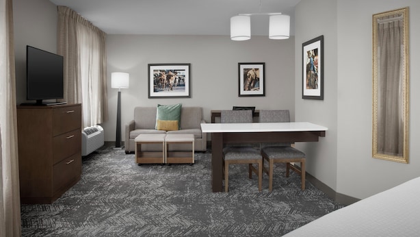 A hotel room with a flat-screen TV, a sofa with cushions, a dining table with chairs, a desk, and a large mirror.