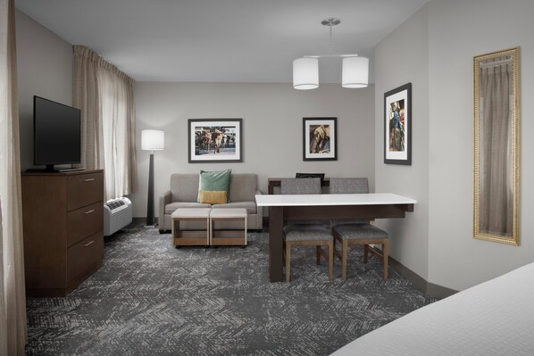 Habitación de hotel con televisión de pantalla plana, un sofá con cojines, una mesa de comedor con sillas, un escritorio y un espejo grande.