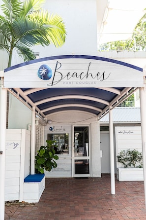 Uma entrada com tema praiano, apresentando uma palmeira, uma placa com a inscrição "Beaches Port Douglas" e uma área de recepção.