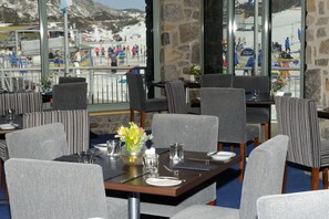 A restaurant with tables set for dining, a vase with flowers, and a view of a ski slope with people in the background.