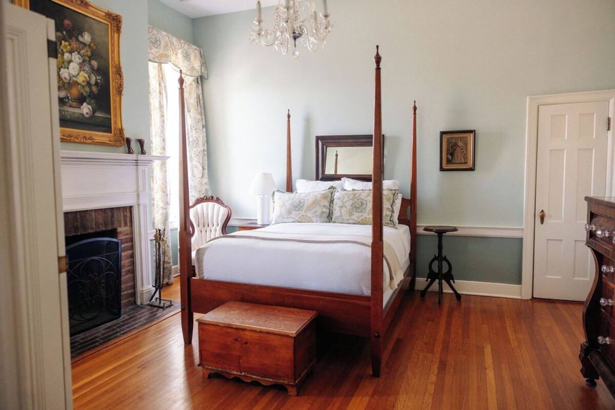 A four-poster bed with a wooden base, a small bedside table, and a nightstand. There is a fireplace with a mantel and a framed floral painting above it.