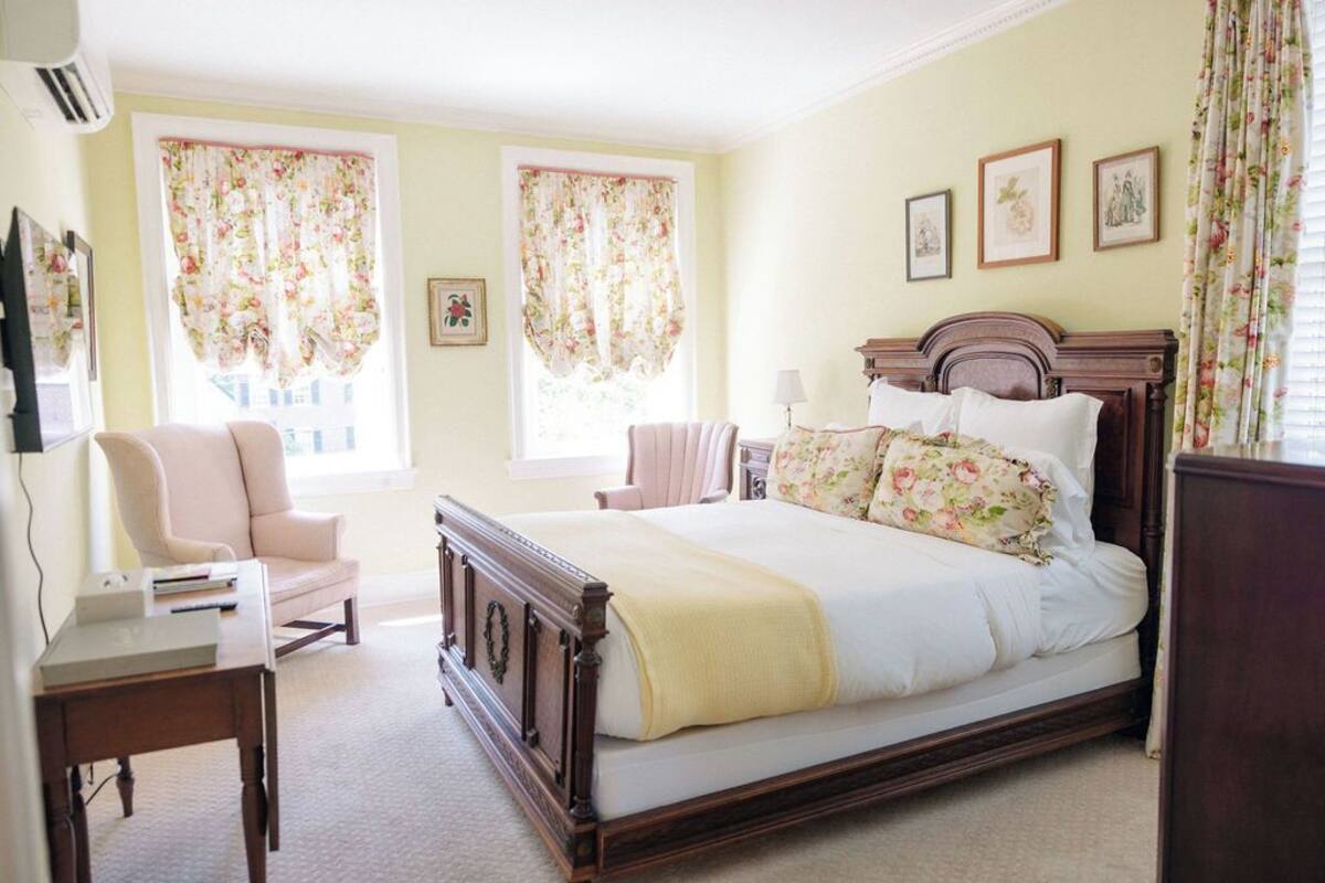 A bedroom with a large bed, two chairs, a desk, and a window with floral curtains.