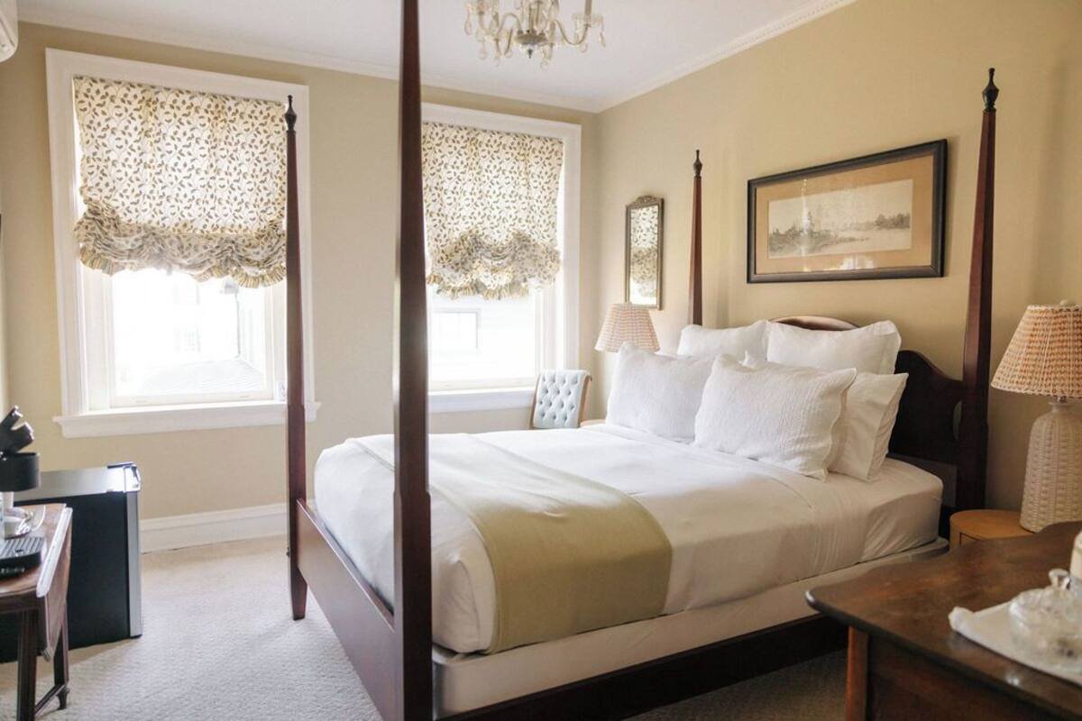 A four-poster bed with white linens, a desk with a lamp, and a framed picture on the wall.