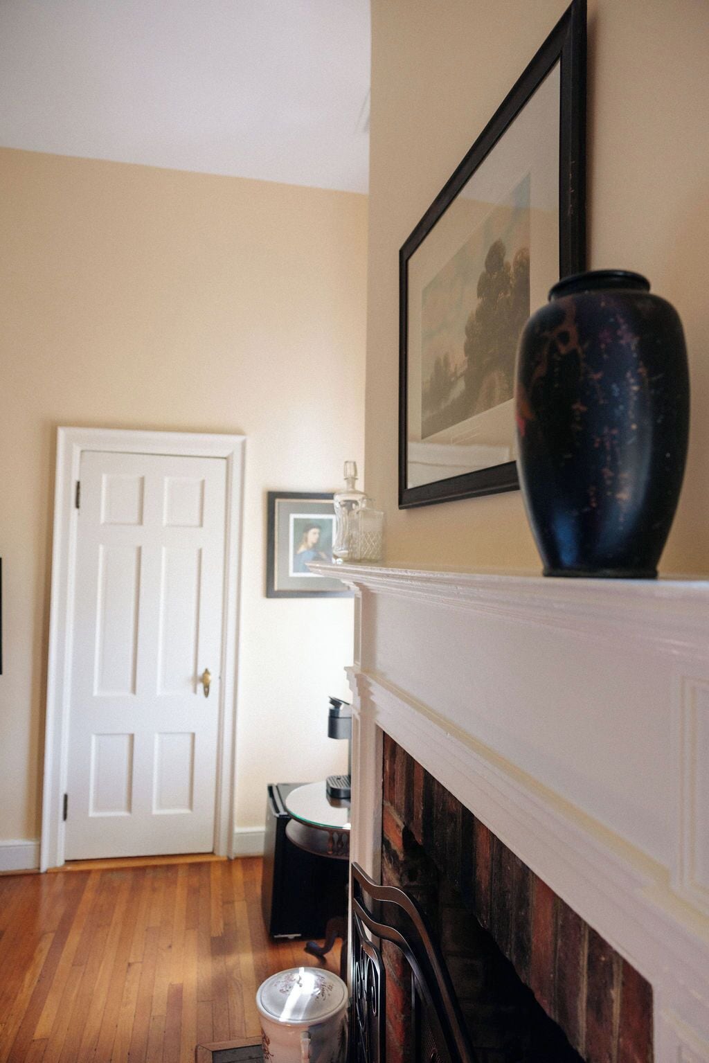 A room with a white door, a fireplace with a black vase, and a framed picture on the wall.