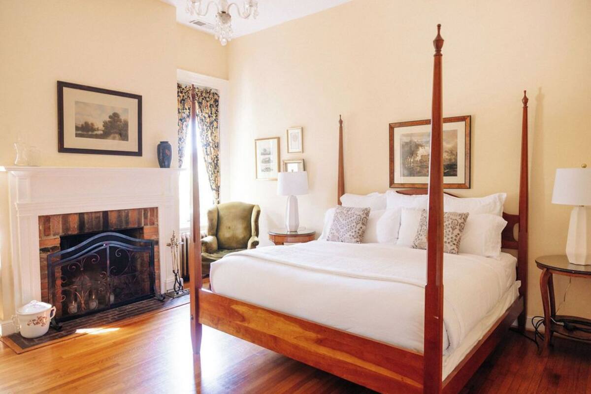 A four-poster bed with a wooden frame, a fireplace with a white mantel, and framed pictures on the walls.