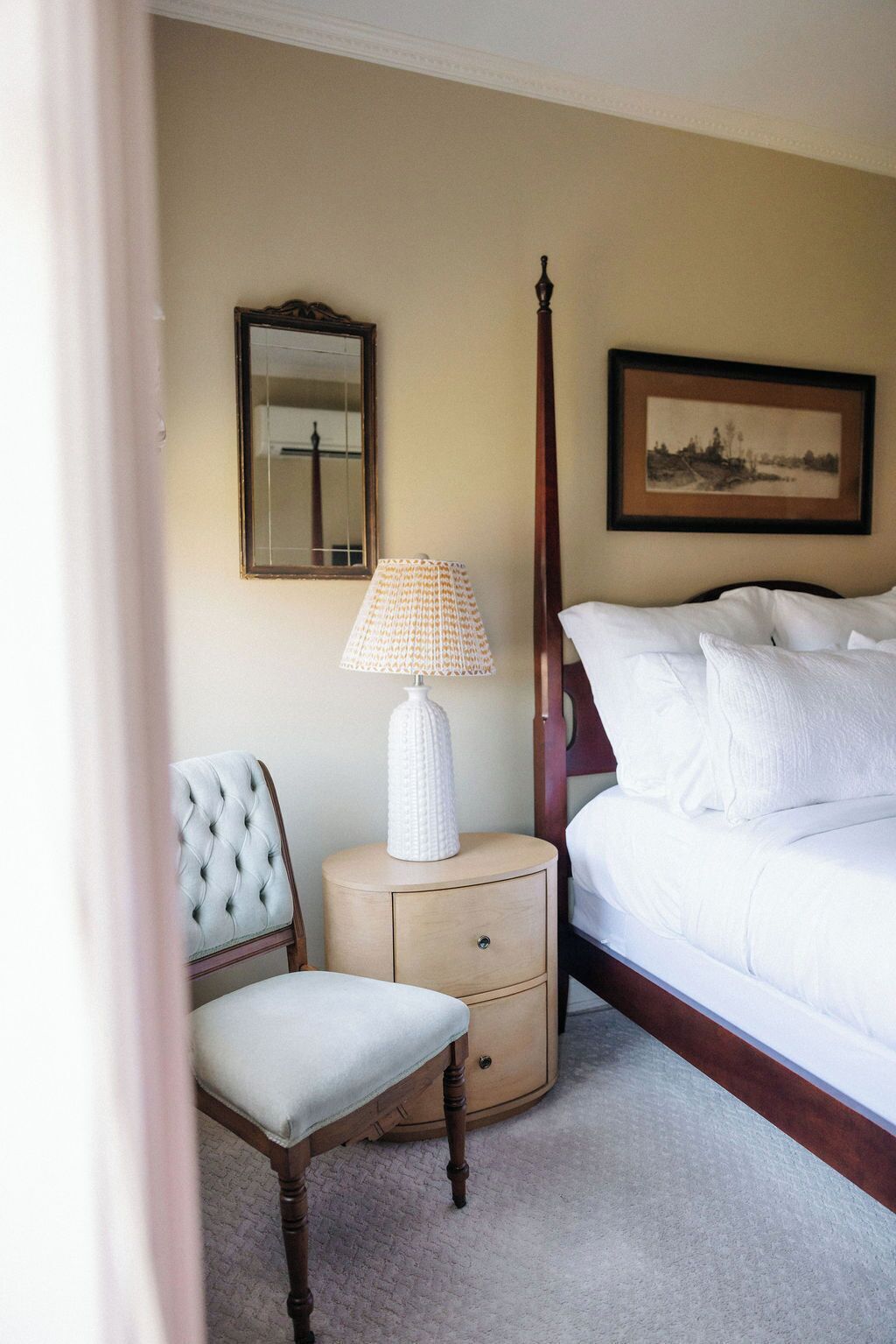 A hotel room with a bed, a nightstand with a lamp, a chair, a mirror, and a framed picture on the wall.