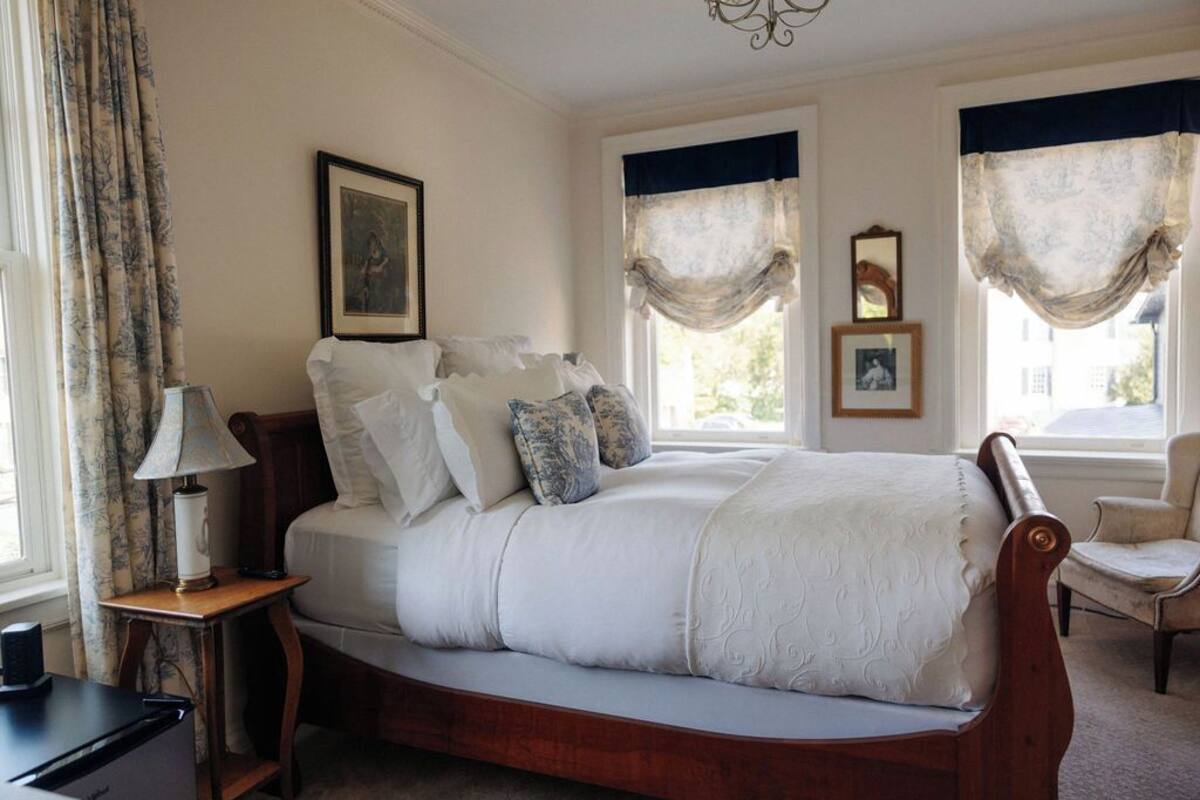 A bedroom with a large bed, a bedside table with a lamp, two windows with curtains, and a chair.