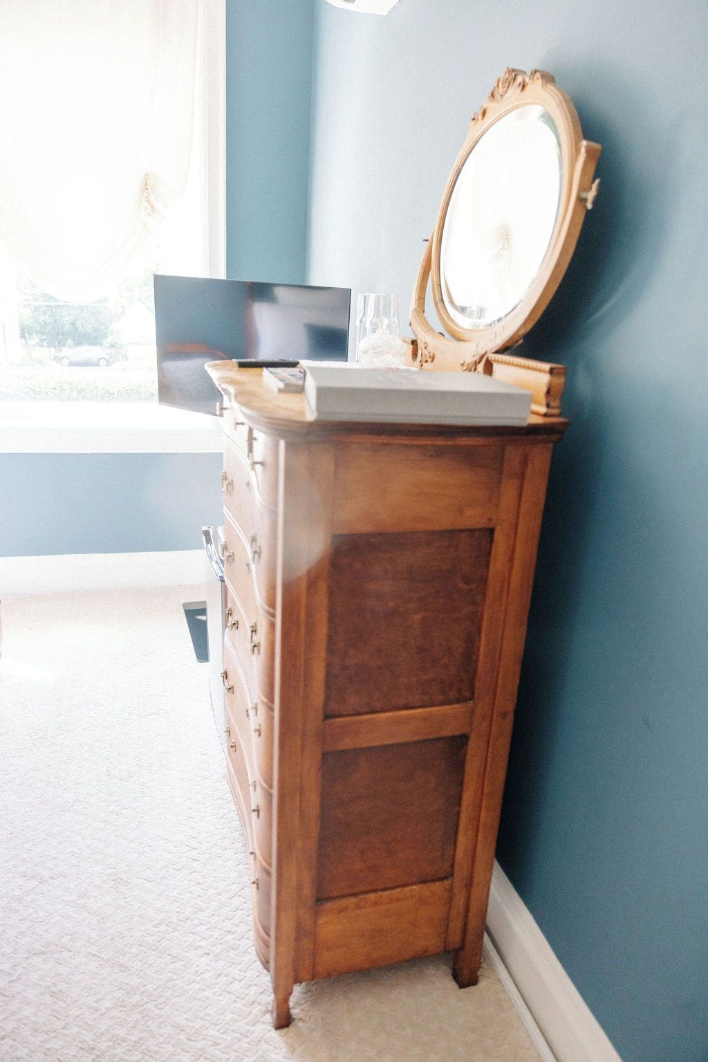 A wooden dresser with a mirror and a laptop in a room with blue walls and a window.