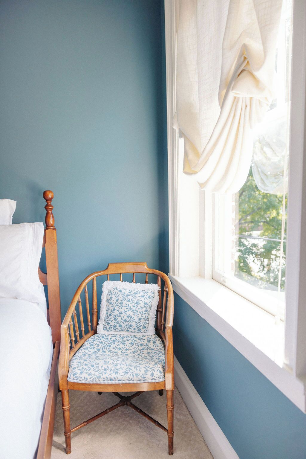 A bedroom with a wooden chair, a bed with white linens, and a window with a white curtain.