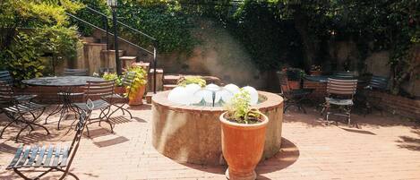 An outdoor patio with brick flooring, metal chairs, a round table, and a central planter with a plant.