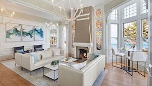 A spacious living room with a fireplace, large windows, and a chandelier. - Green Gables Inn, A Four Sisters Inn (Pacific Grove)