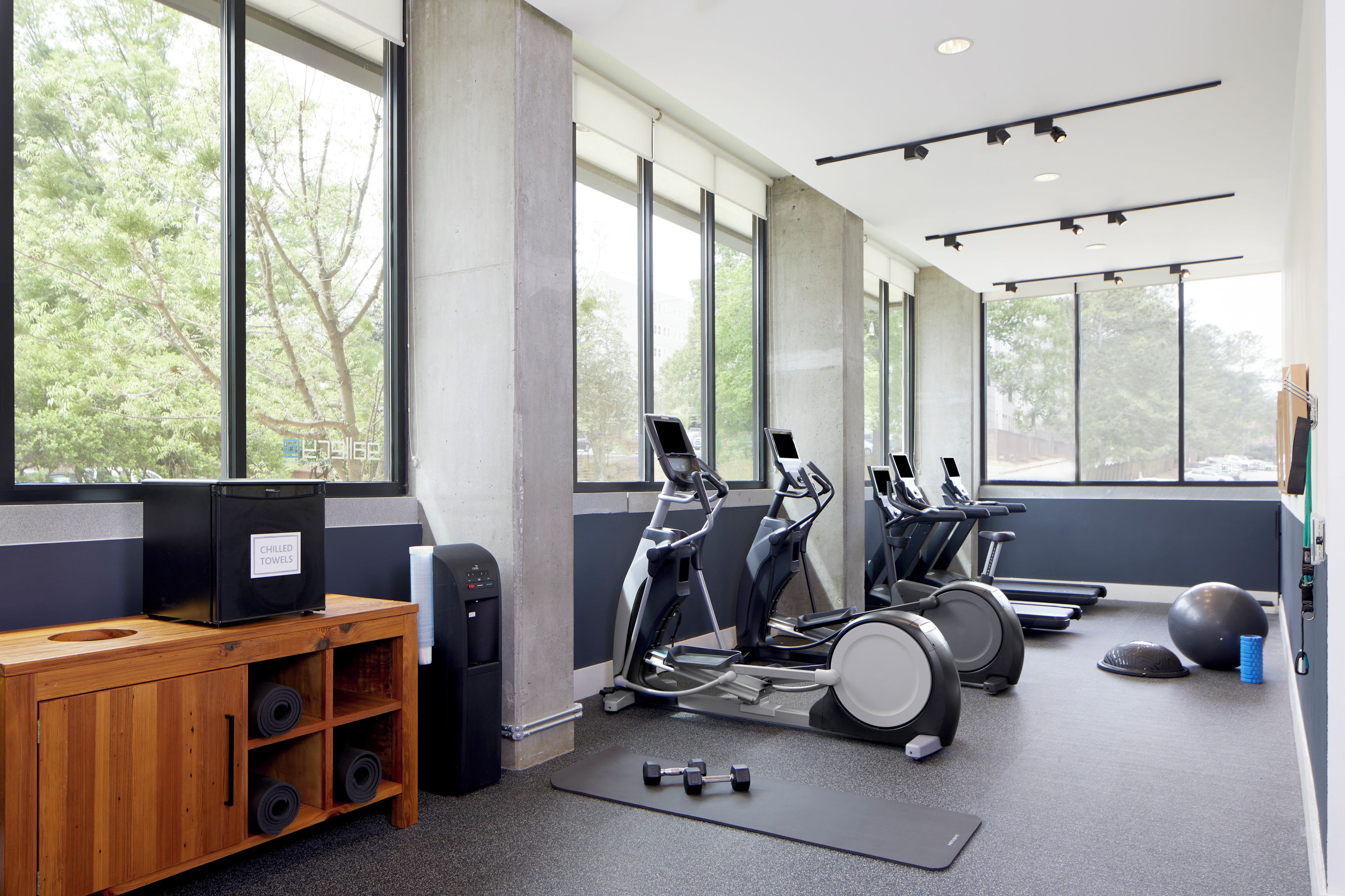 A gym with exercise equipment, a wooden cabinet with towels, and large windows.