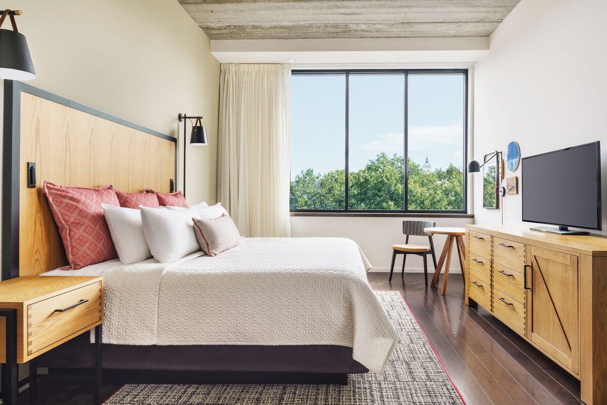 A modern hotel room with a large bed, a television, a desk, and a view of greenery outside.