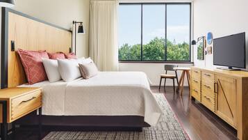 A modern bedroom with a large bed, a TV, a wooden dresser, and a window with a view of trees.