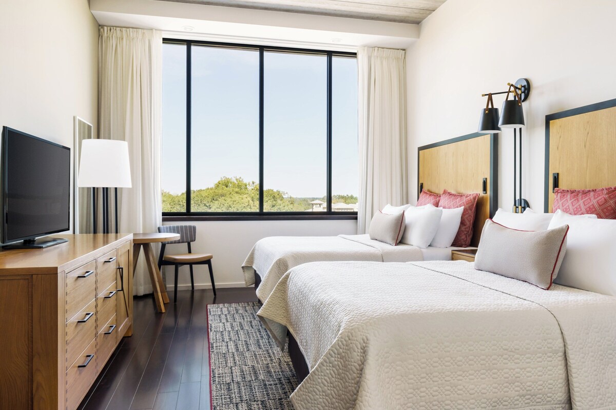 A hotel room with two beds, a television, a desk, and a view of the outdoors.