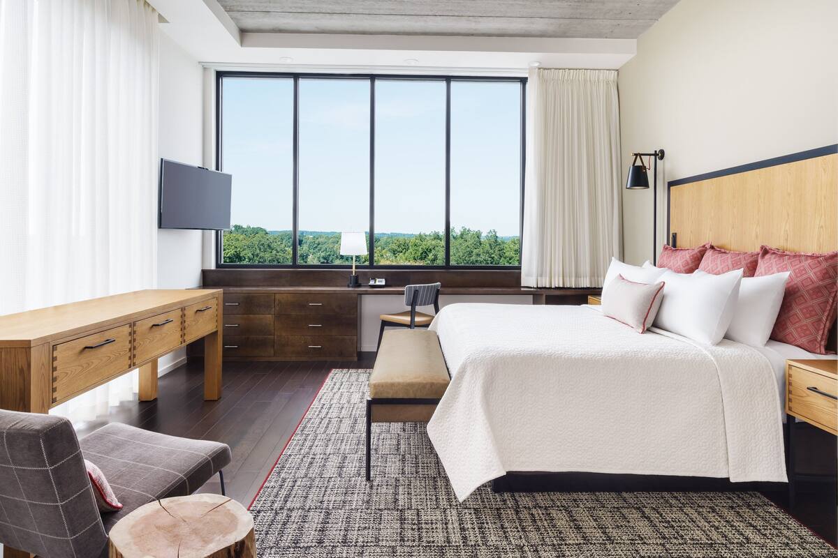 A modern hotel room with a large window, a bed with white bedding, a desk with drawers, a chair, and a small table.