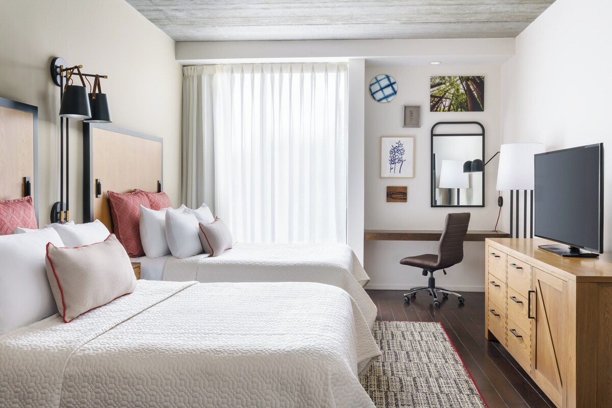 A hotel room with two beds, a desk, a television, and a window with curtains.