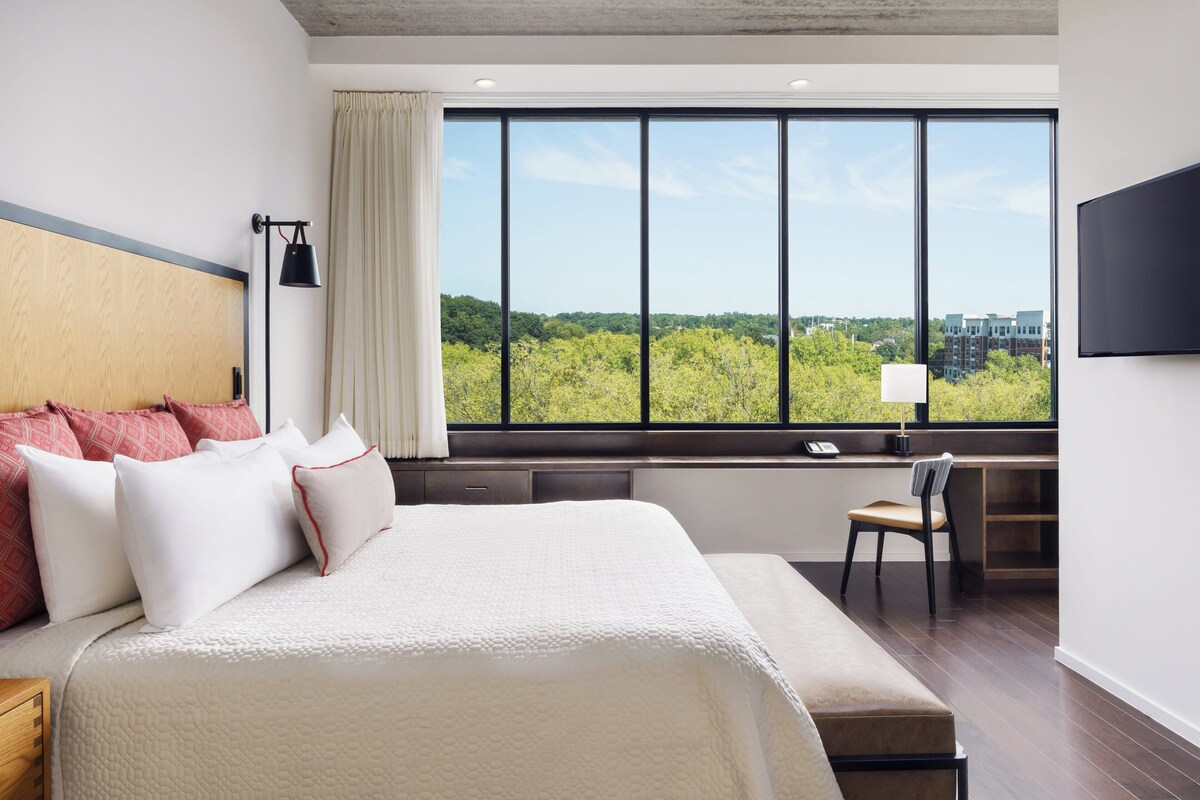 A hotel room with a large bed, a desk, and a view of greenery outside.