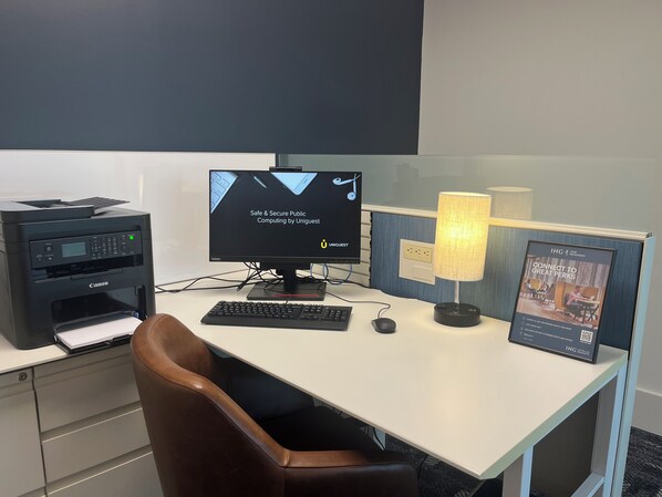 A hotel workspace with a Canon printer, a computer monitor, a keyboard, a mouse, a desk lamp, and a framed poster.