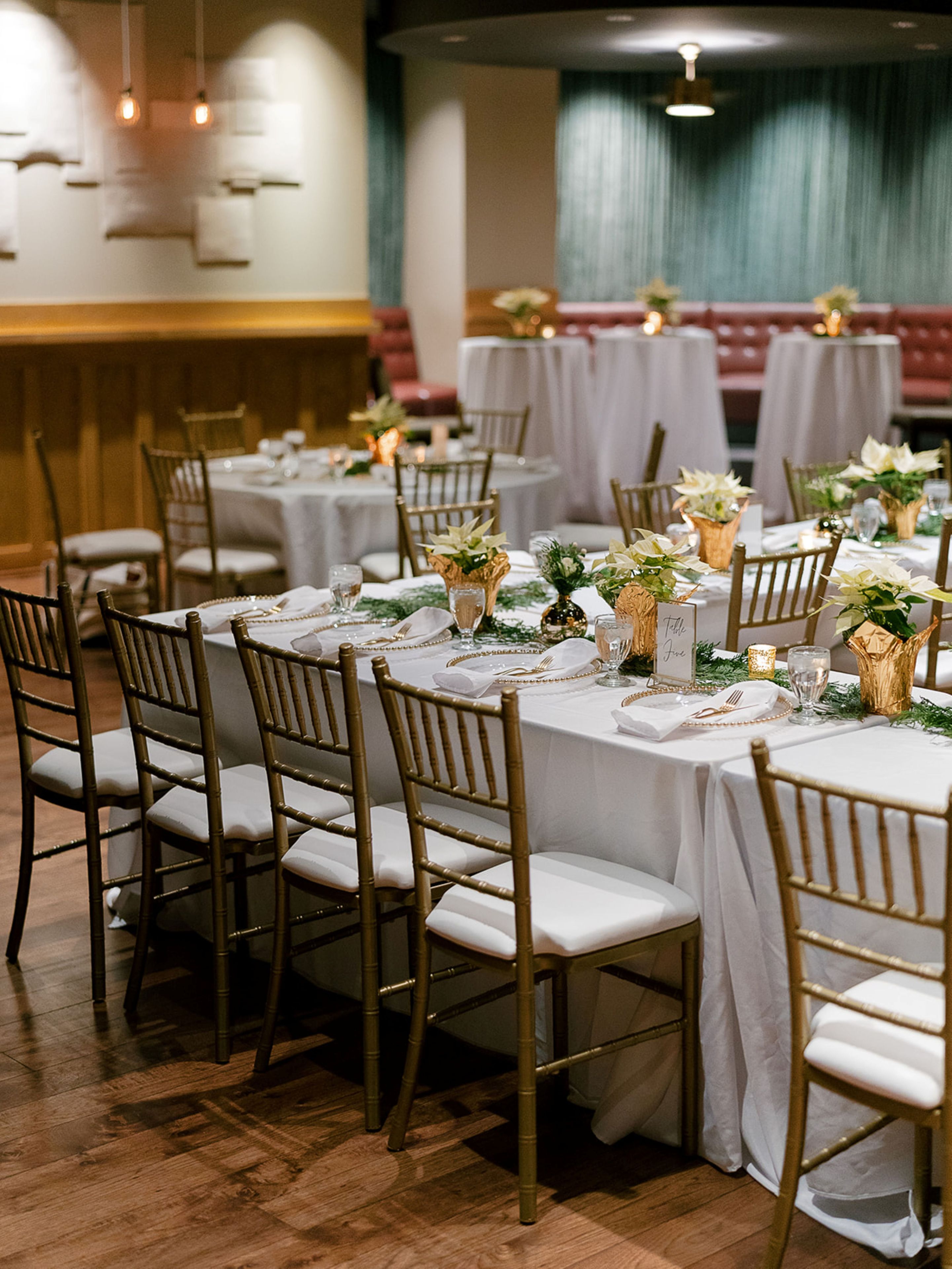 Une salle de banquet avec des tables rondes dressées pour le repas, chacune étant recouverte d’une nappe blanche, agrémentée de chaises dorées et d’arrangements floraux.
