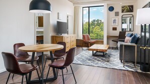 A modern living room with a wooden dining table, leather chairs, a sofa, and a large window offering a view of trees. - Hotel Indigo Athens University Area by IHG (Athens)