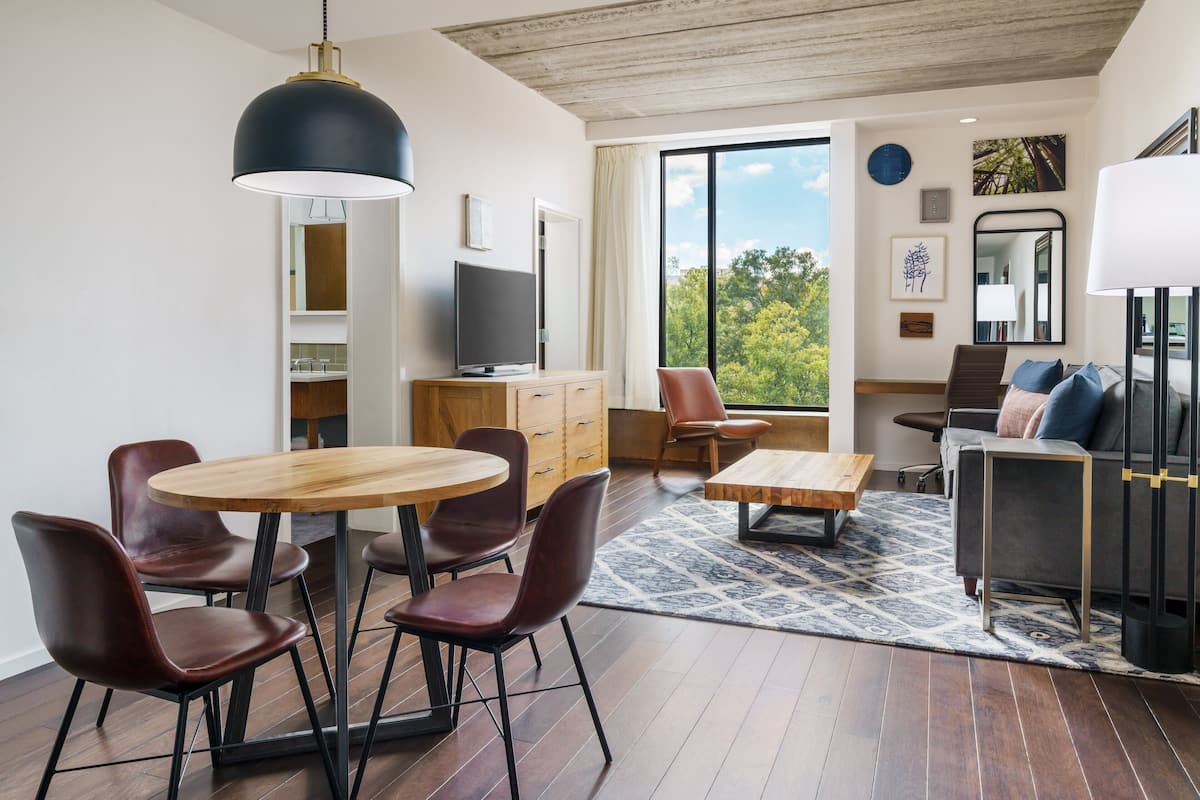 Een moderne woonkamer met een houten eettafel, leren stoelen, een bank en een groot raam met uitzicht op bomen.