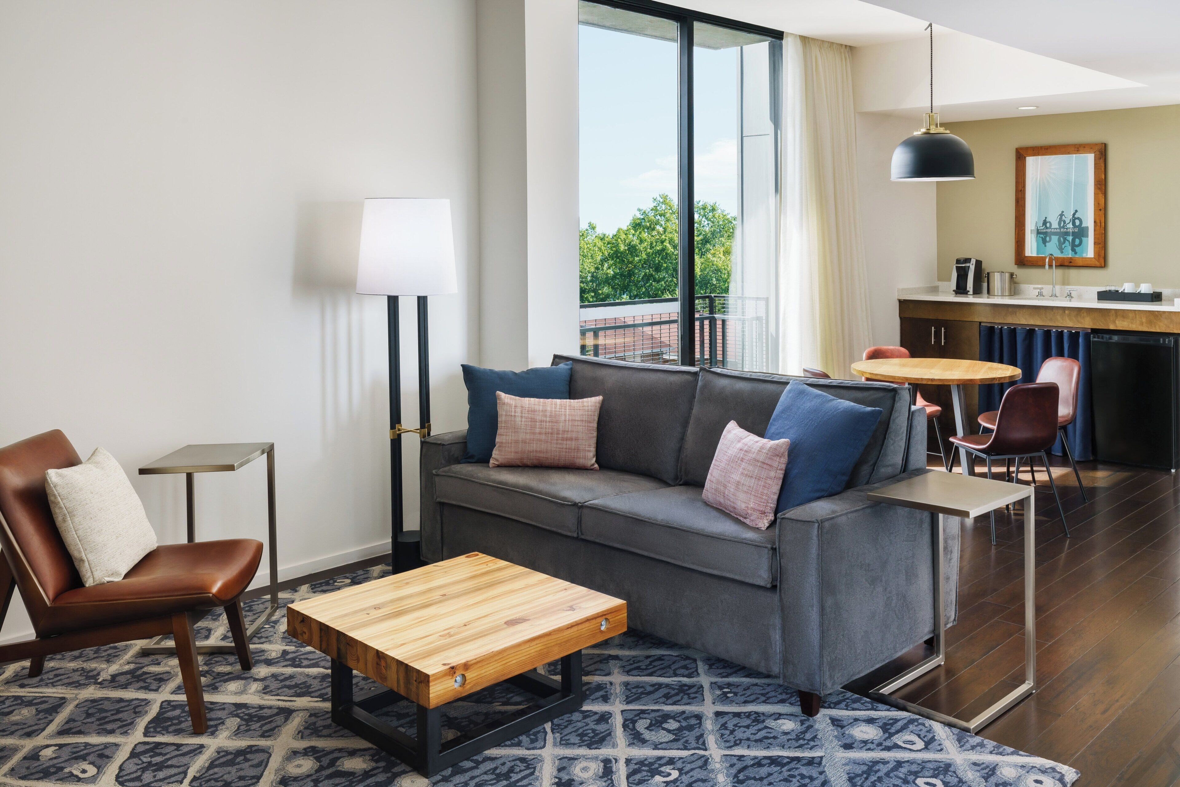 A modern hotel room with a grey sofa, a wooden coffee table, a patterned rug, and a view of greenery through large windows.