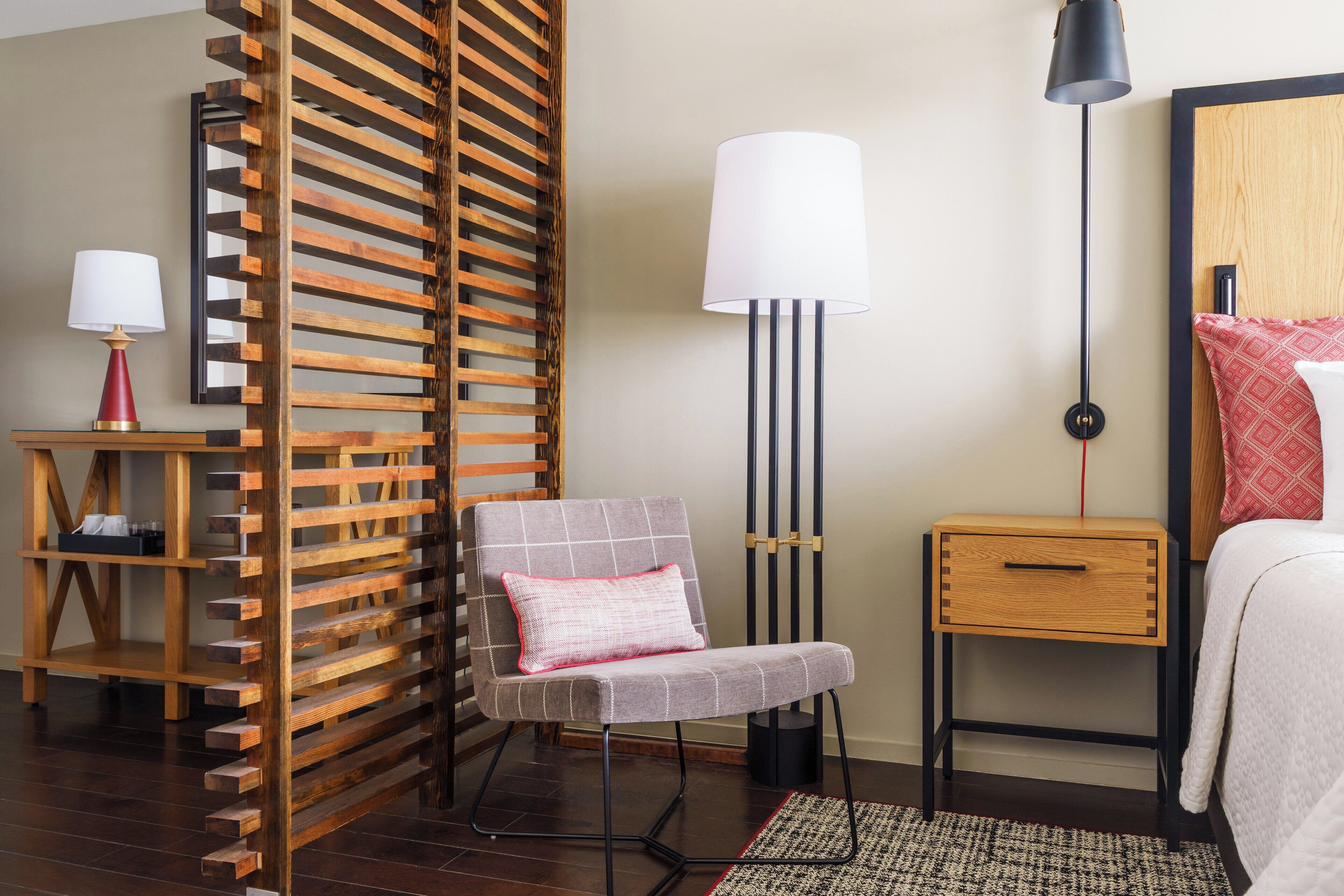 A hotel room with a wooden slatted partition, a chair with a cushion, a bedside table with a lamp, a bedside drawer, and a bed with a patterned pillow.