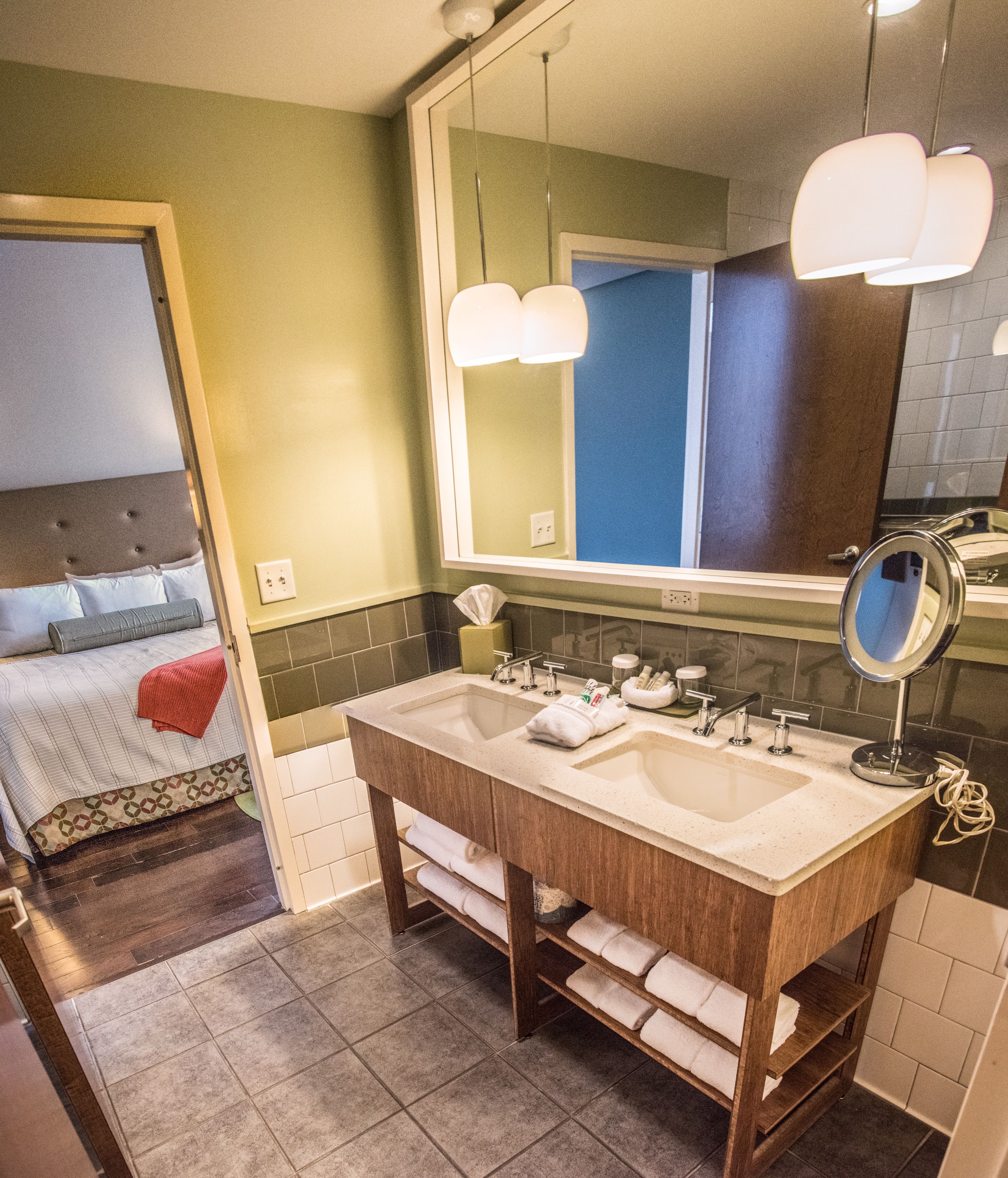 A hotel bathroom with a double sink, a large mirror, and a bed with a headboard and pillows.