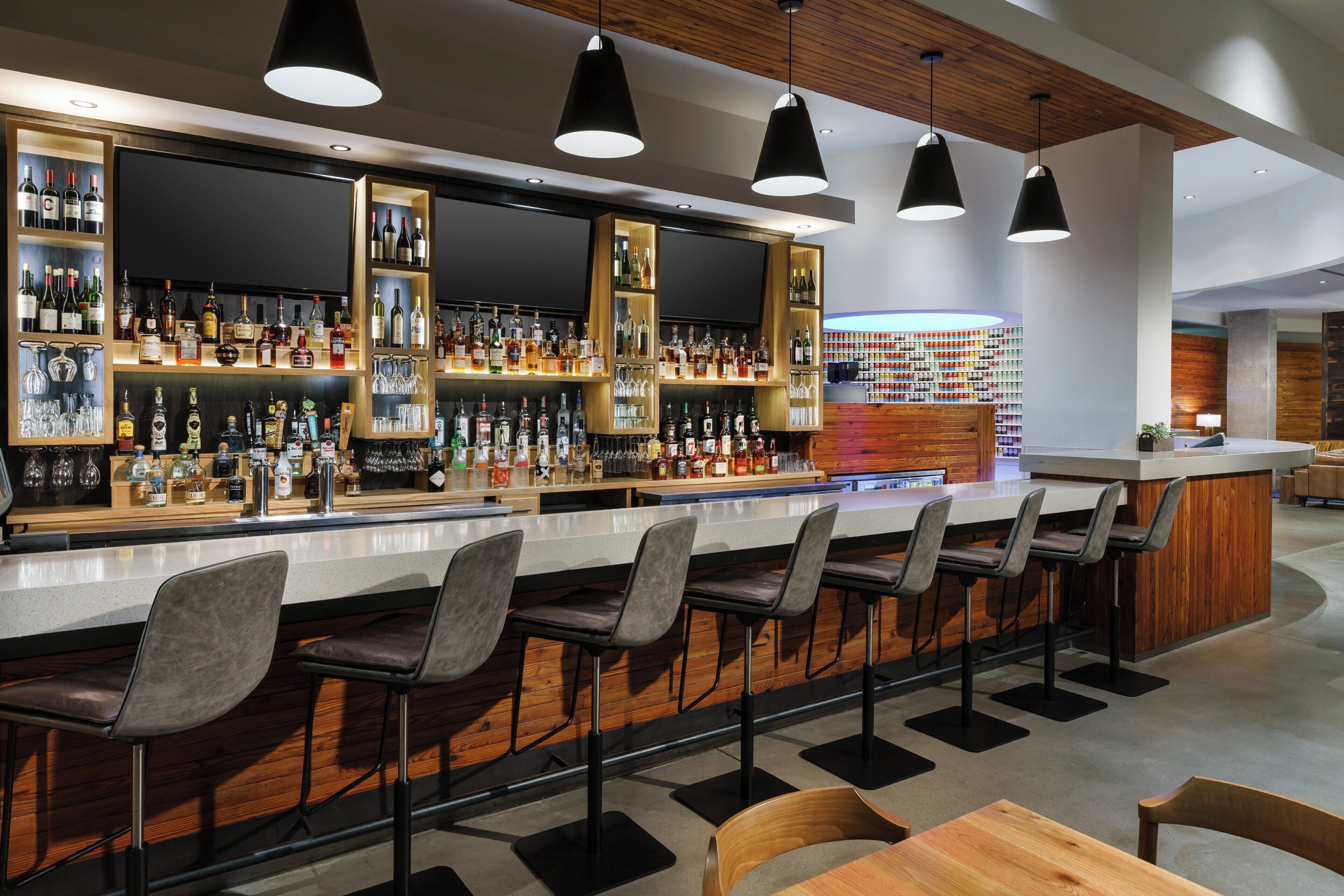 Uma área de bar com várias prateleiras de garrafas de bebidas, banquetas e iluminação pendente.