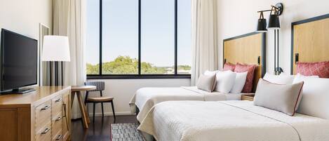 A hotel room with two beds, a TV, a desk, and a large window with a view of greenery.