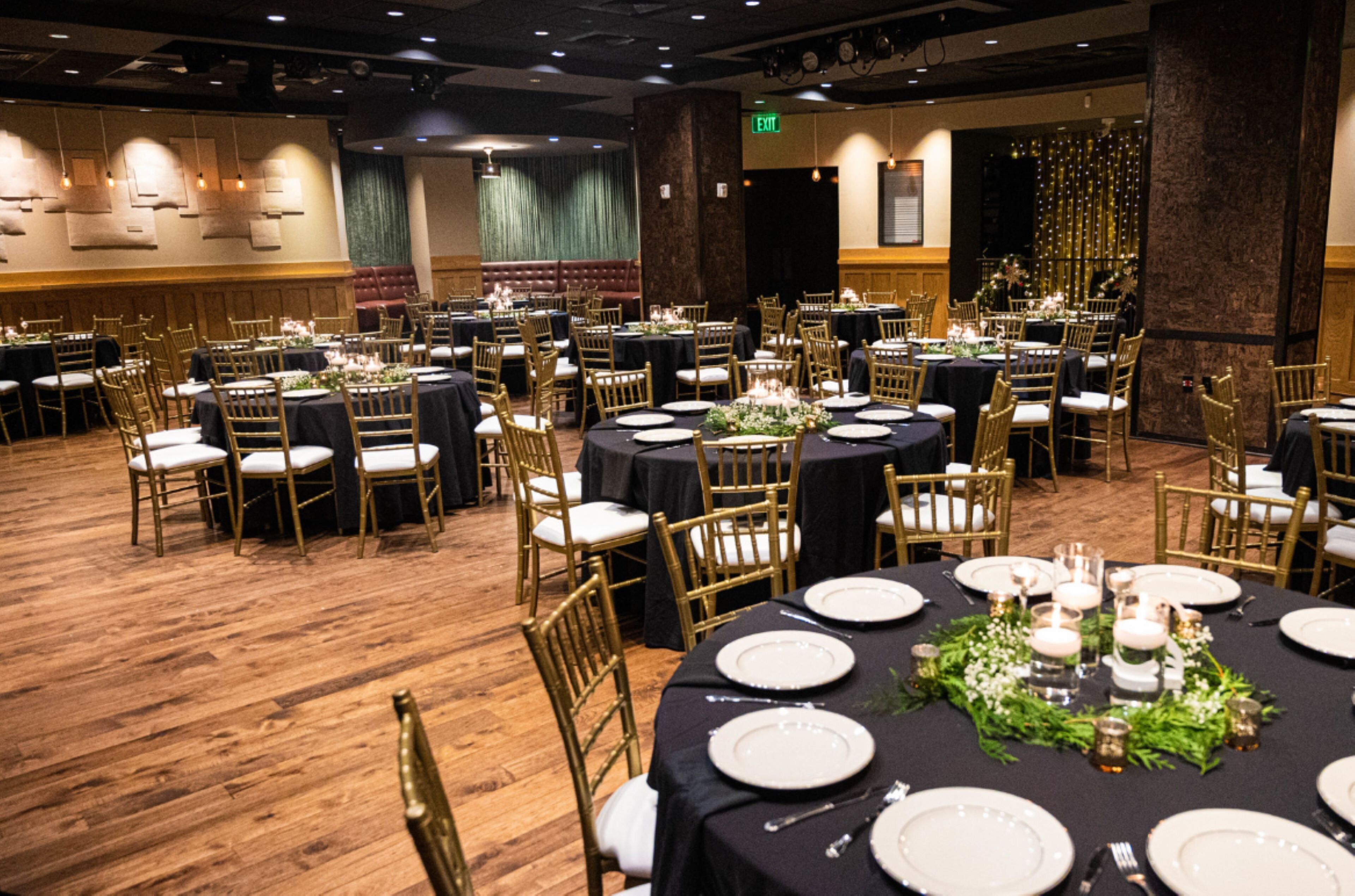Une salle de banquet avec des tables rondes dressées pour le repas, des chaises dorées et un plancher en bois.