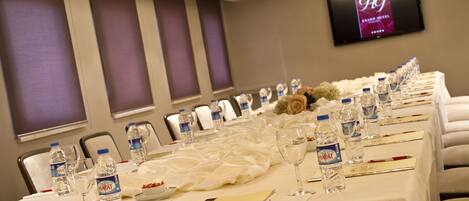 Un salón de conferencias con una larga mesa dispuesta para una reunión, equipado con botellas de agua, copas de vino y centros de mesa florales.