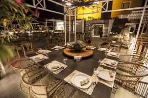 An outdoor dining area with round tables and wicker chairs, surrounded by a white structure with plants.