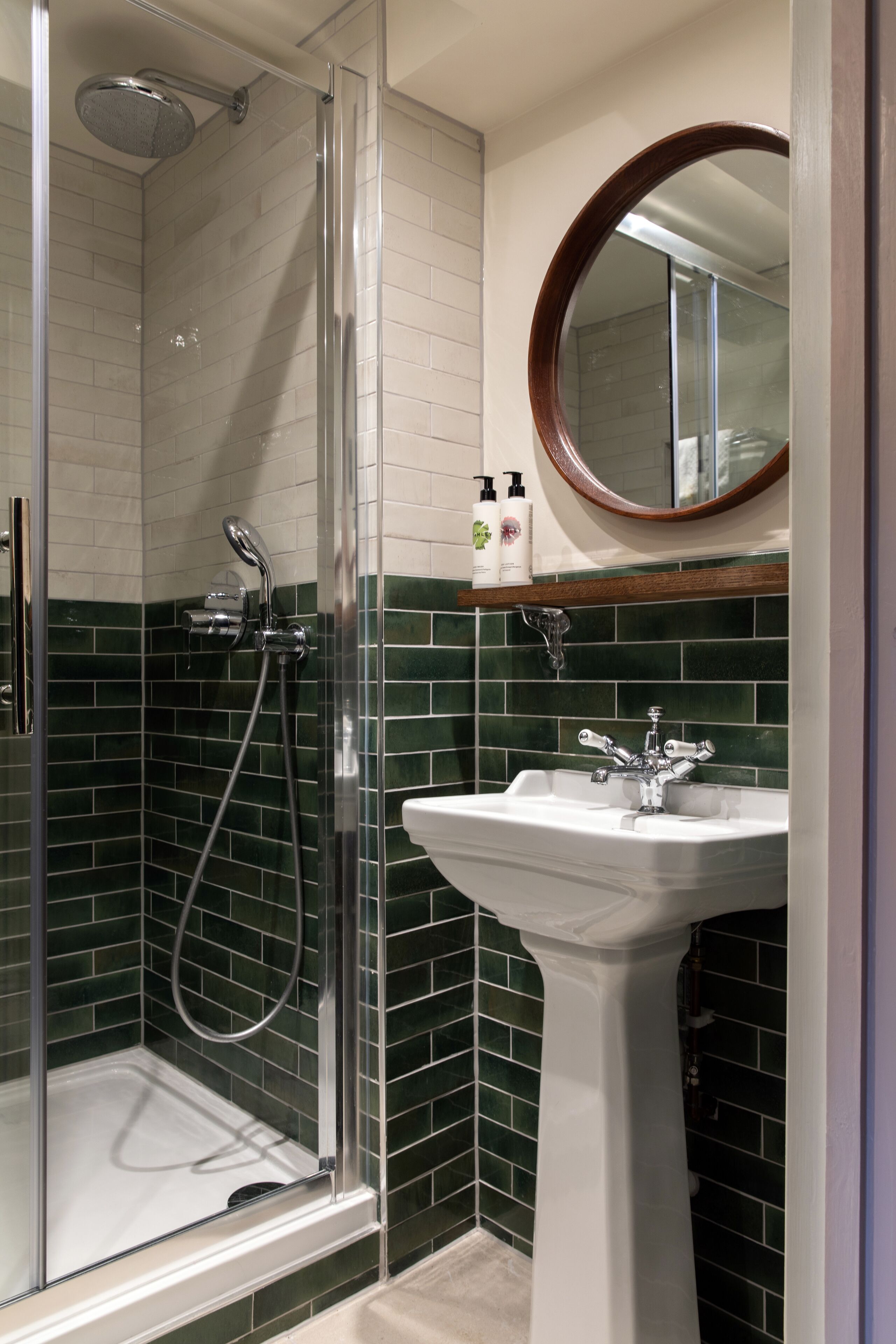Een badkamer met een douche met glazen wand, een wit zuilwastafeltje en een spiegel boven de wastafel.