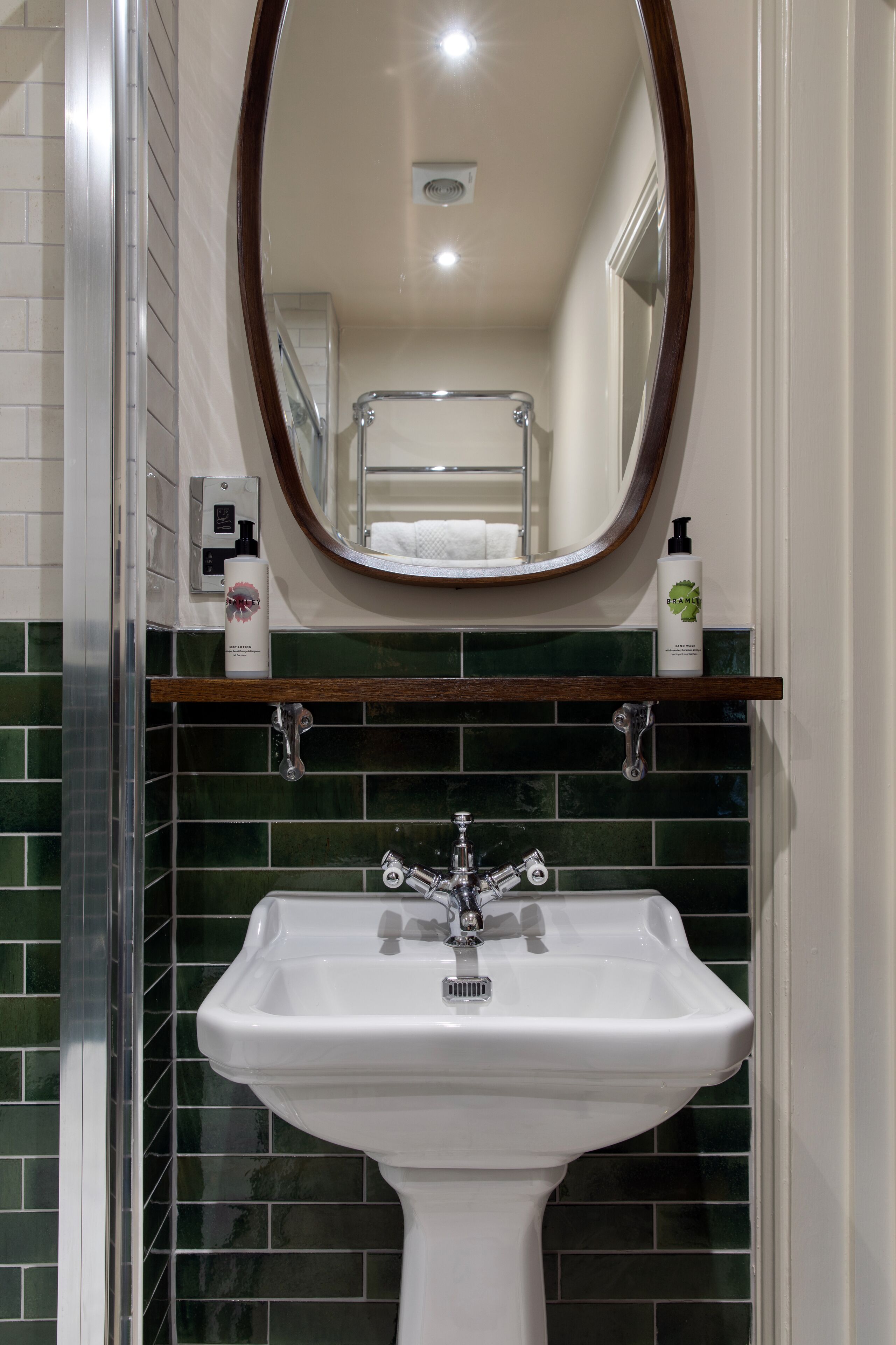 A bathroom with a white pedestal sink, green tiled backsplash, a wooden shelf with toiletries, and a mirror above the sink.