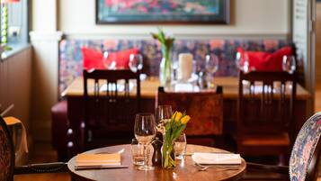 Una mesa de comedor dispuesta con copas de vino, un jarrón con flores amarillas y una vela. Hay sillas alrededor de la mesa, y un banco con cojines rojos al fondo.