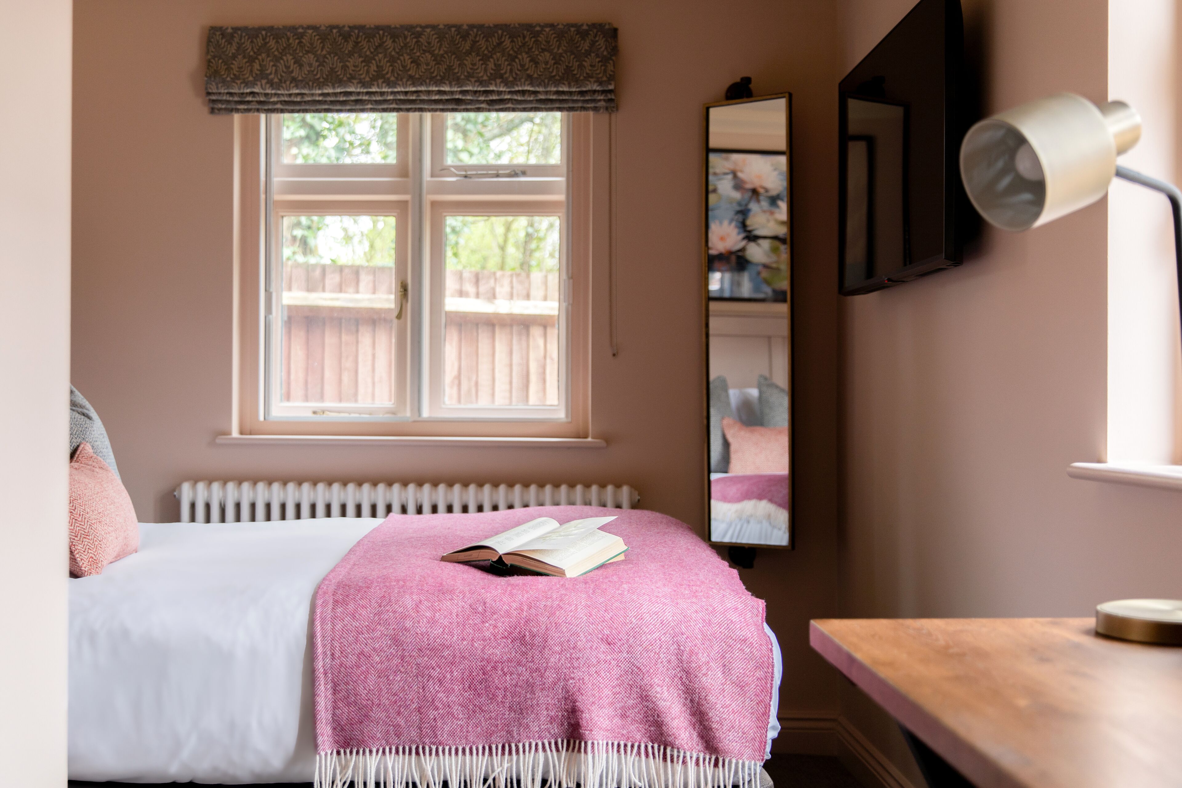 A bedroom with a bed, a bedside table, a window with a view of the outside, and a wall-mounted mirror.