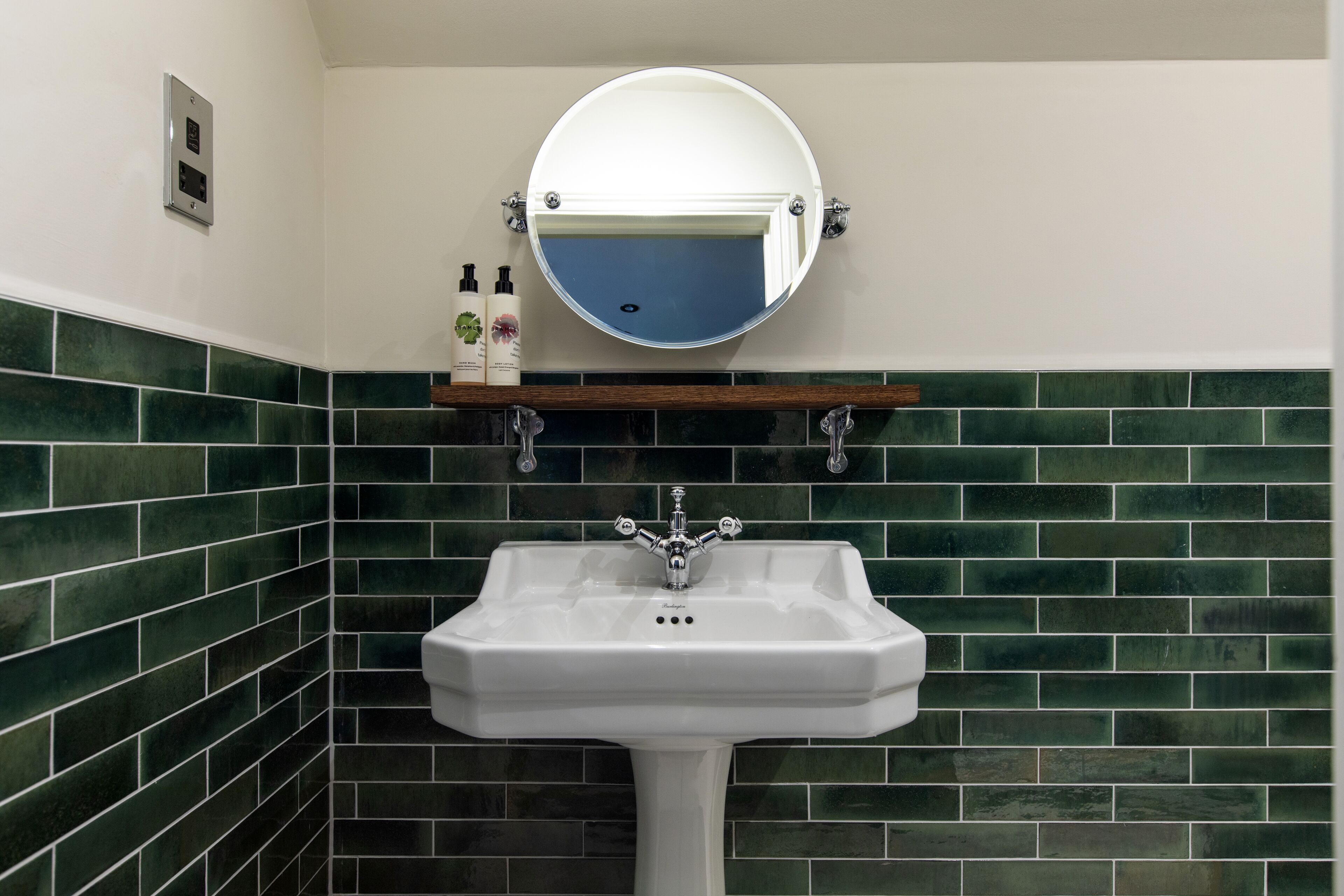 Een badkamer met een wit wastafeltje op een voetstuk, een ronde spiegel en groene betegelde wanden.