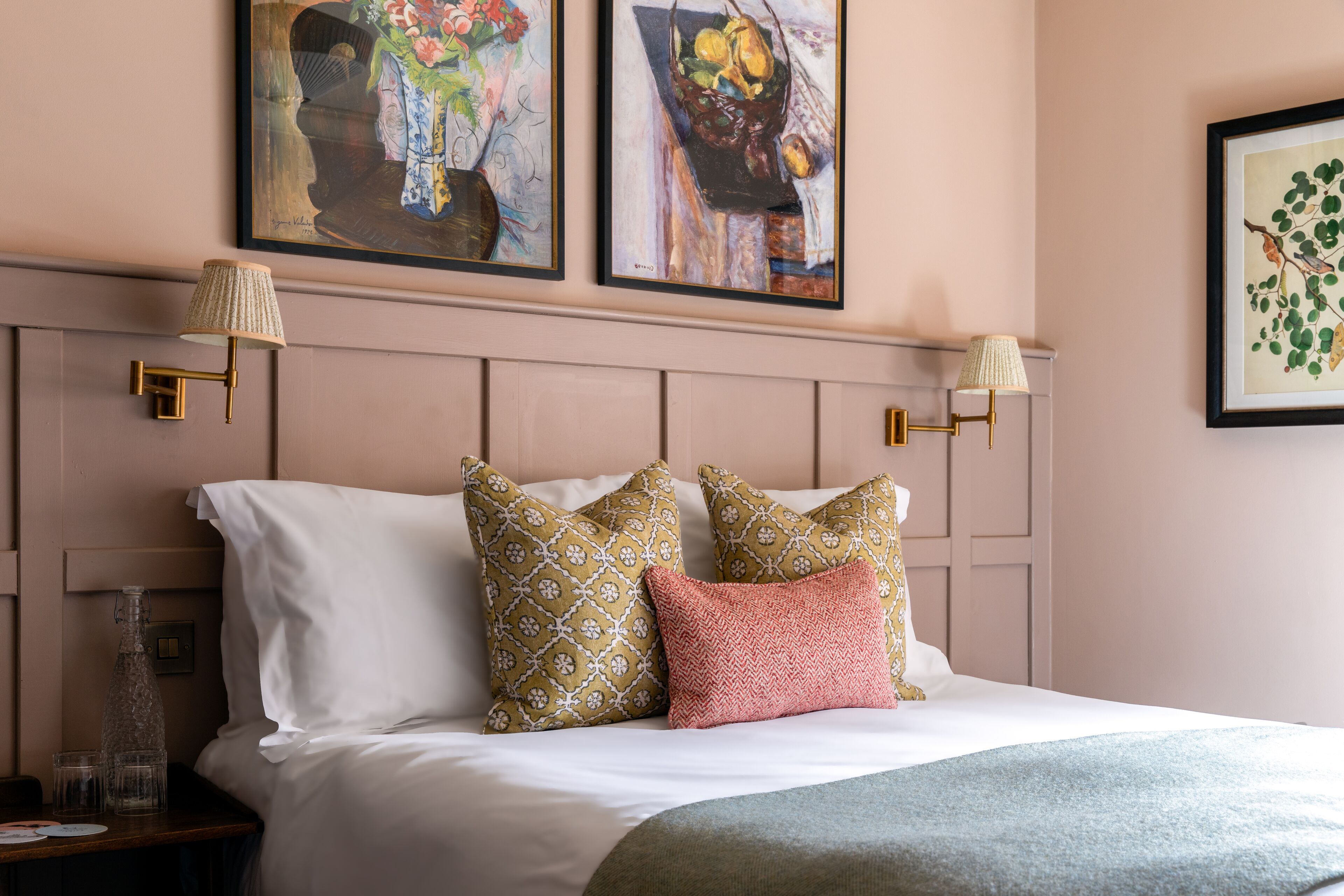 A neatly made bed with patterned pillows, a bedside table with glasses and a bottle, and wall-mounted lamps.