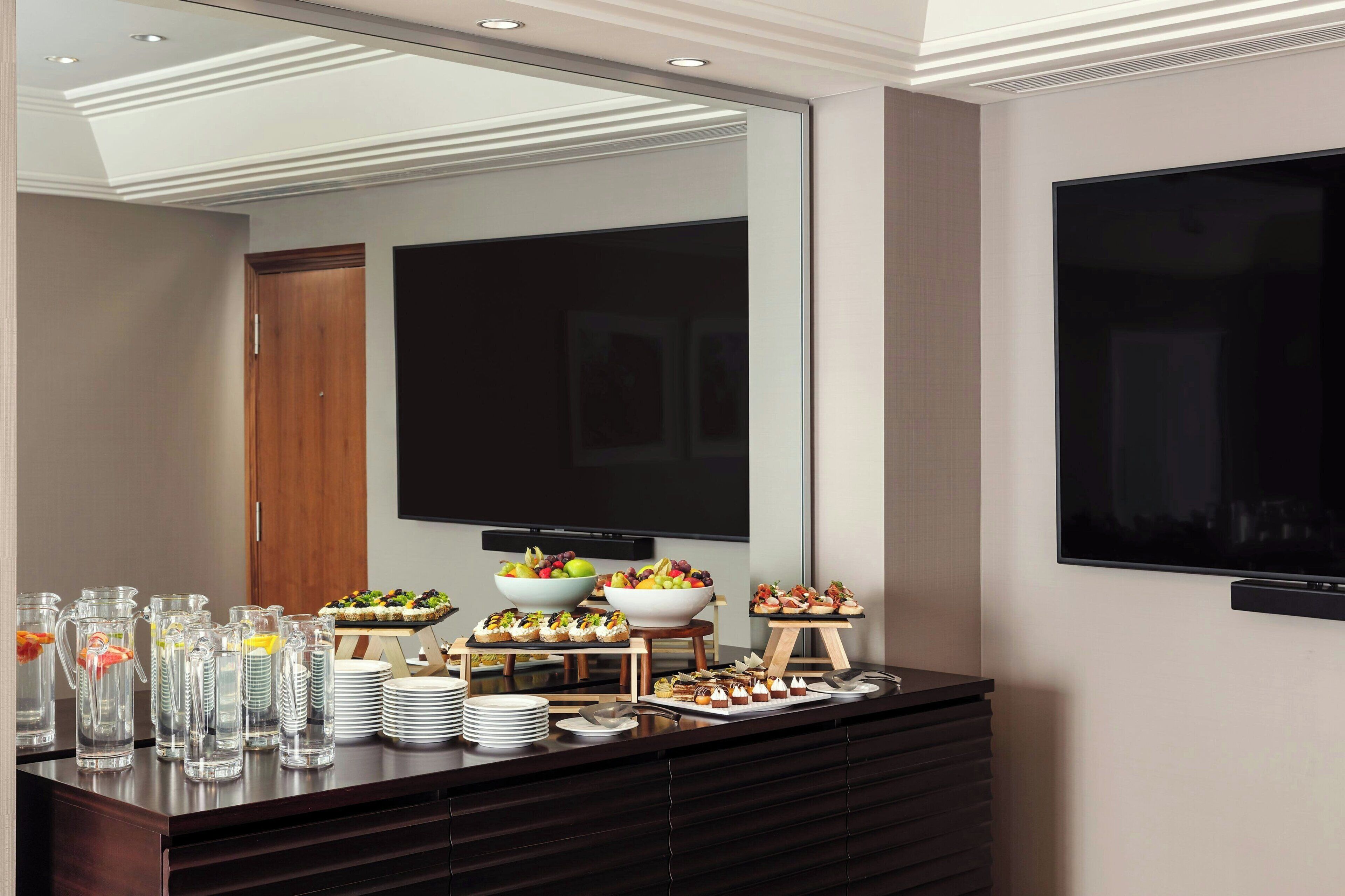 A buffet setup with various food items and beverages, a large mirror, and a flat-screen TV mounted on the wall.