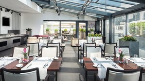 Een modern restaurant met een glazen dak, netjes opgestelde tafels en potplanten.