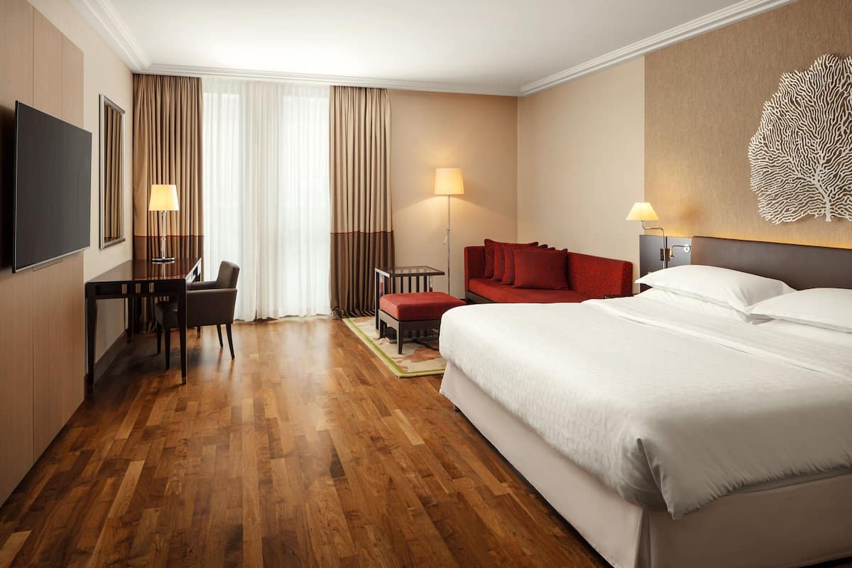 A hotel room with a large bed, a desk with a chair, a red sofa, a TV, and a wall-mounted artwork.