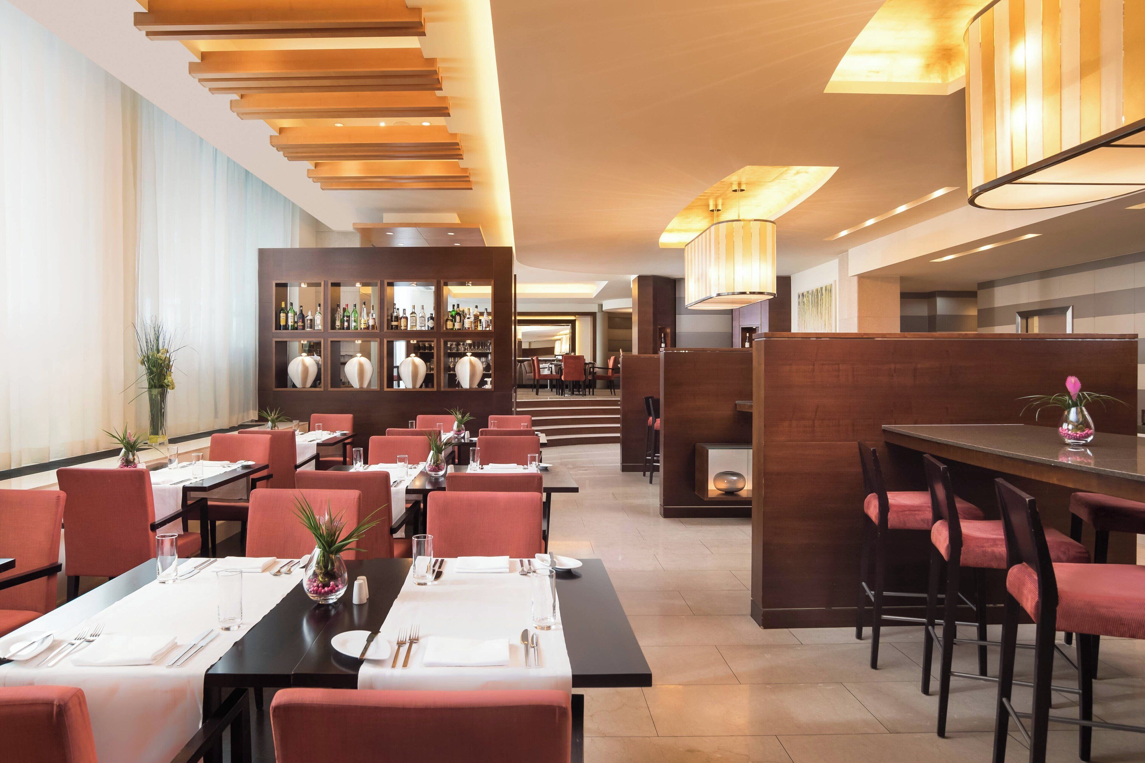 A modern dining area with tables set for two, featuring red upholstered chairs and a central bar area with a variety of bottles.