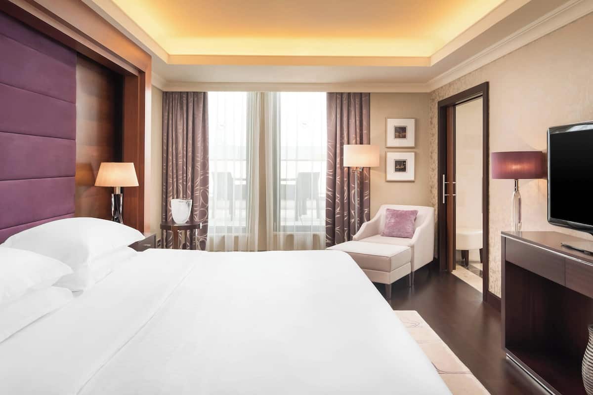 A hotel room with a large bed, a desk, a chair, a TV, and a window with curtains.