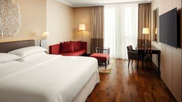 Quarto de hotel com uma cama grande, um sofá vermelho, uma mesa de trabalho com uma cadeira e uma televisão.