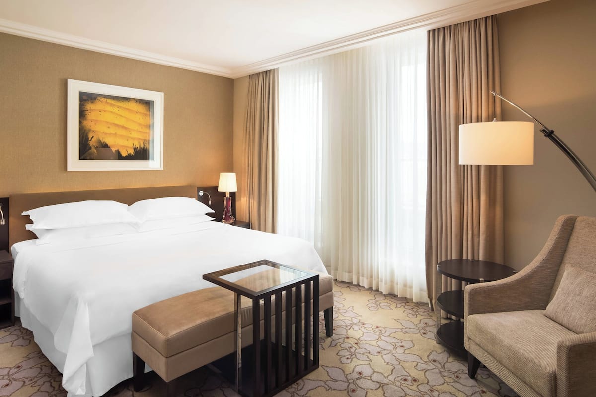 A hotel room with a large bed, a bedside table with a lamp, a chair, and a painting on the wall.